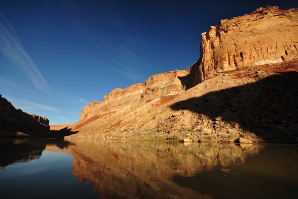 The rocky shore against the background of a clear sky is reflected in the waters of the river