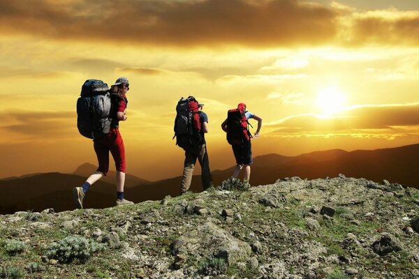 Tres excursionistas con mochilas en la cima de la montaña admiran el sol del atardecer