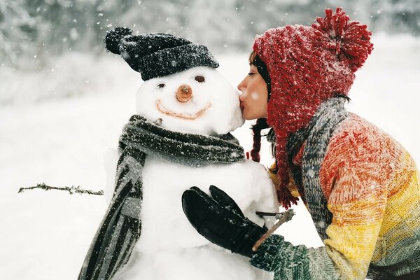 A girl kisses a snowman in a hat