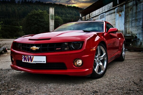 A red Chevrolet stands on a dirt road against the backdrop of mountains