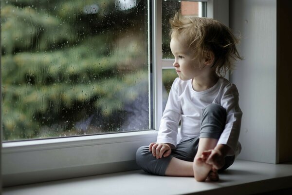 Bebê menina senta-se no peitoril da janela e olha pela janela