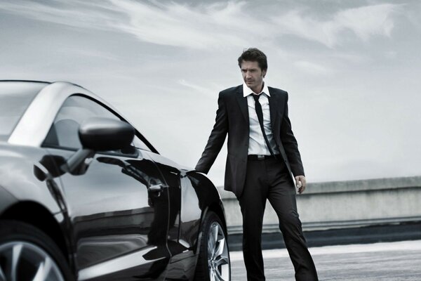 A man in a suit stands near a black car