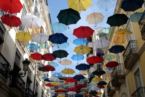 Multicolored umbrellas hang over the street between buildings against a blue sky background