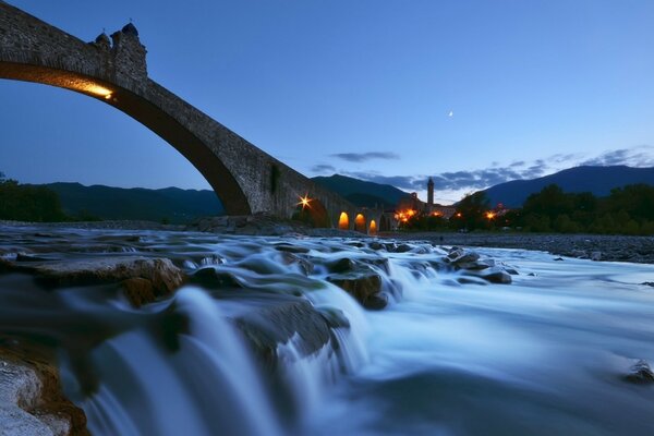 Eine fließende, steinige Flussschwelle in Italien mit einer mit gelben Lichtern beleuchteten Brücke