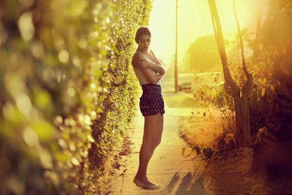 A girl in shorts and ballet flats hugs herself against the background of a garden path