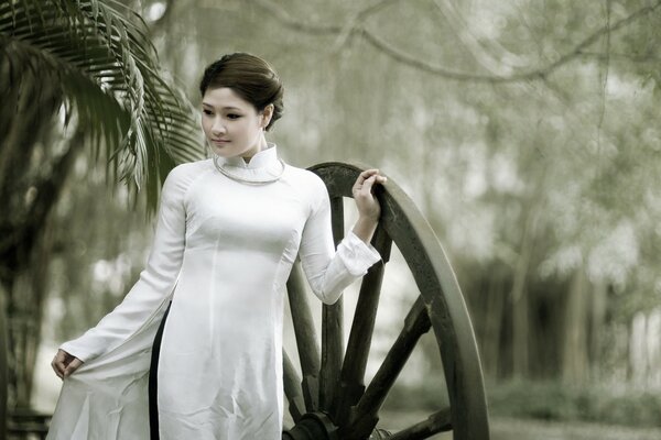 فتاة آسيوية في ثوب أبيض مغلق تقع يدها على عجلة خشبية كبيرة