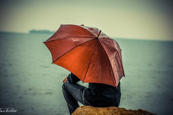 Ein Mann sitzt auf einem Stein am Meer unter einem roten Regenschirm