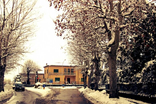 Foto de un camino cubierto de nieve y un edificio amarillo detrás de los árboles