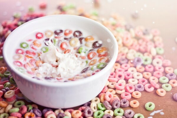 Eine weiße Schüssel mit Milch gefülltem Essen auf einem Tisch zwischen zerbröckeltem, buntem Getreide in Form von kleinen Kreisen