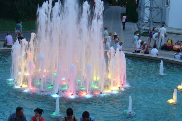 An einem Sommerabend sitzen Menschen am mit verschiedenen Farben beleuchteten Brunnen