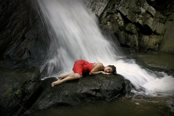 Das Mädchen liegt in einem roten Kleid auf einem Stein neben einem Wasserfall