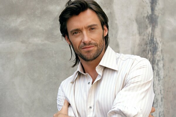 Actor Hugh Jackman stands in a white shirt against a gray wall