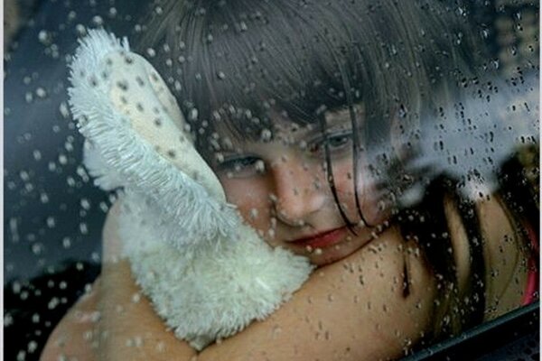 A child with a toy looks out the window with raindrops