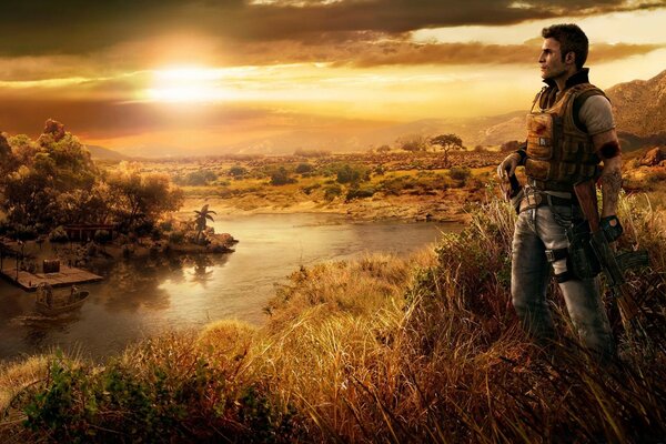 Герой игры Farcry 4 в полной боевой амуниции стоит на высоком берегу реки по колено в траве на фоне закатного неба