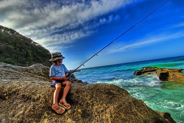 A boy in a panama hat with a fishing rod sits on the stone shore of the blue sea during the day