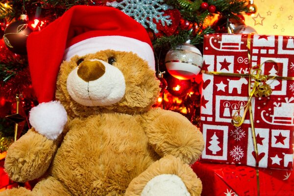 A teddy bear in a Christmas hat on the background of a Christmas tree and a gift