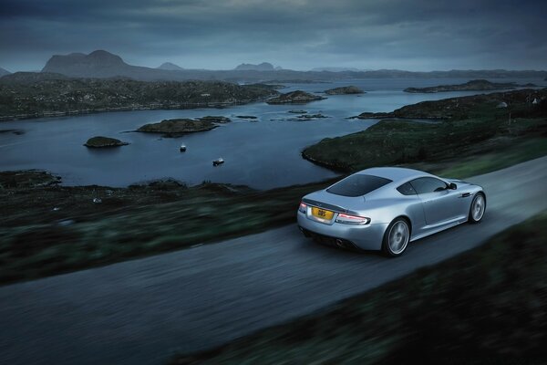 Aston Martin silver color rides along the lake on a country road