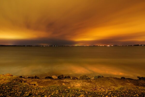 On the shore of a yellow lake under a yellow sky at sunset, and on the other side a glowing city