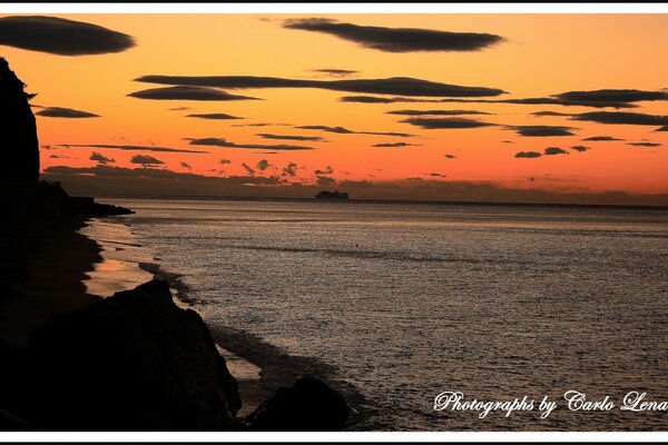 Puesta de sol roja, costa rocosa, mar gris con un barco en la distancia