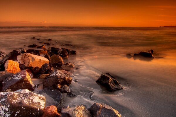 Large boulders in the sand go into the sea water at a yellow-red sunset