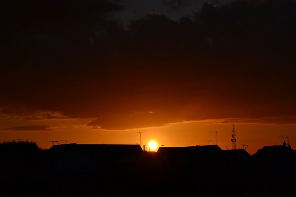 The sunset is hidden behind the dark roofs of houses in a bright orange sky