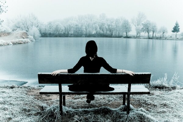 A lonely girl is sitting on a bench near the lake