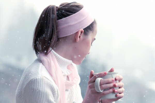 A girl with a pink headband holds a mug in her hands against a background of fine snow